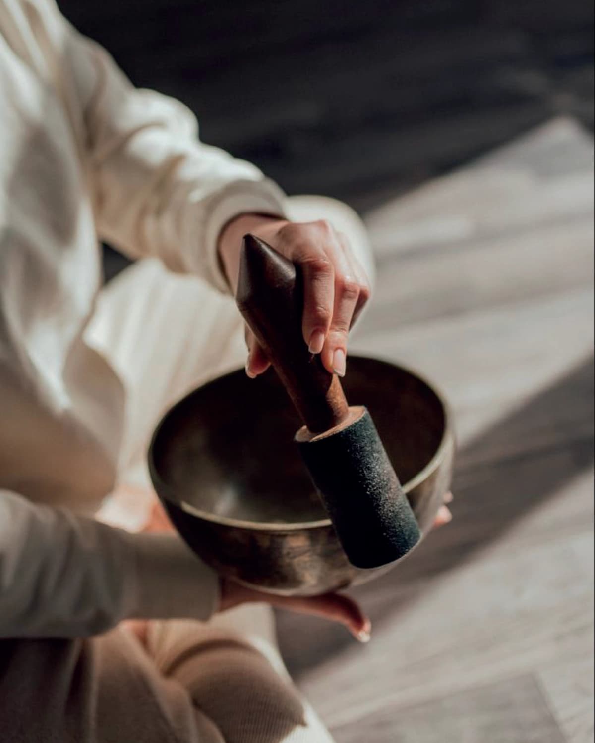 Overhead view of a person dressed in white, seated on a wooden floor, sounding a metal singing bowl with a long-handled mallet