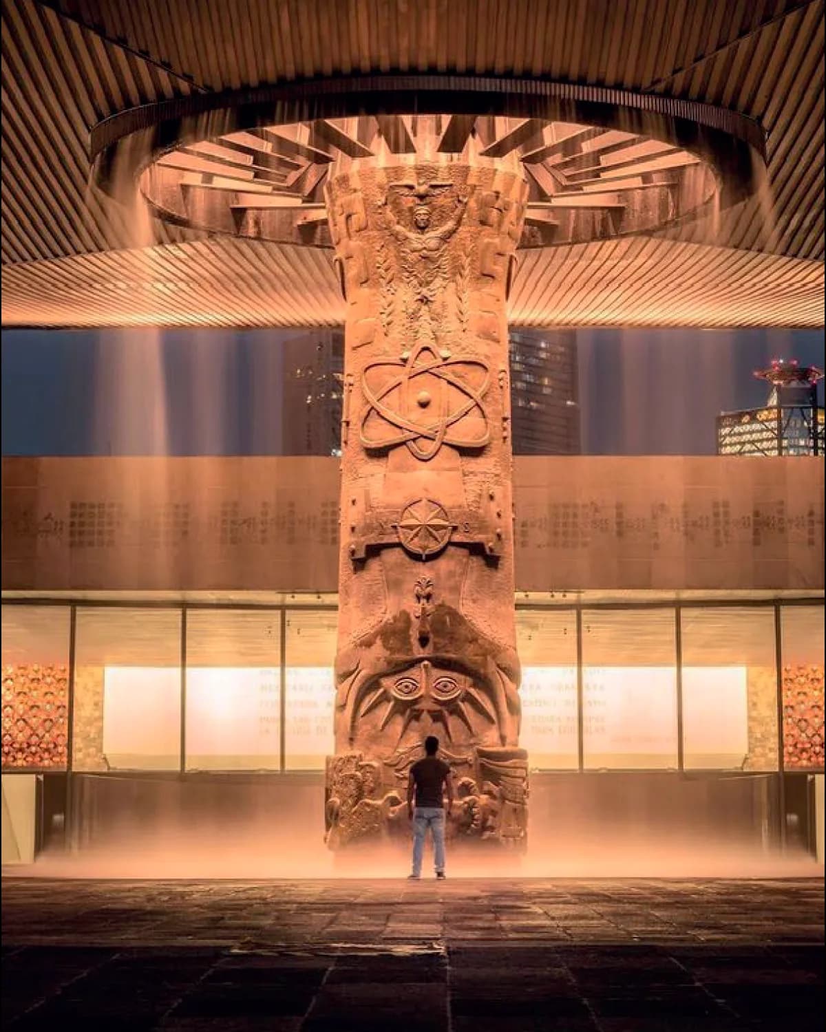 The umbrella fountain of the National Museum of Anthropology lit at night: the carved column with its curtain of water falling from the circular canopy, and a person standing at its base