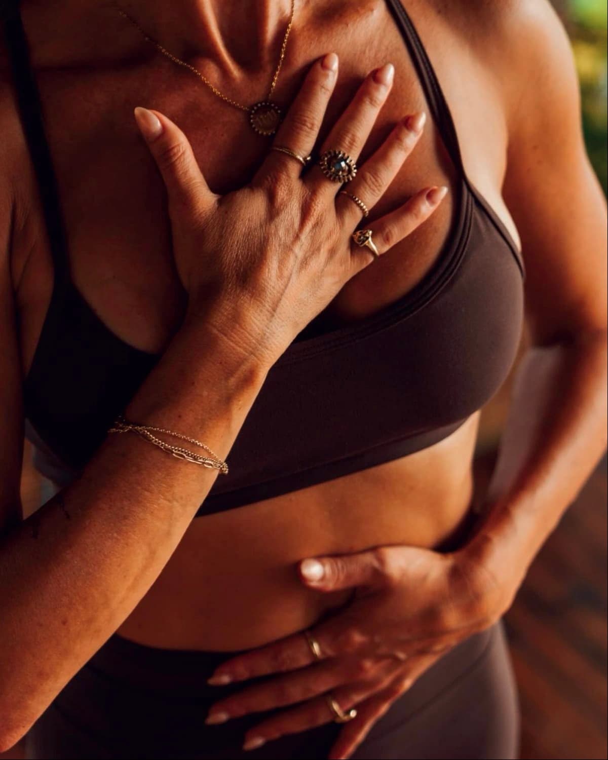 Close-up of a woman's torso in a brown sports top, one hand on her chest and the other on her belly, wearing gold rings and bracelets