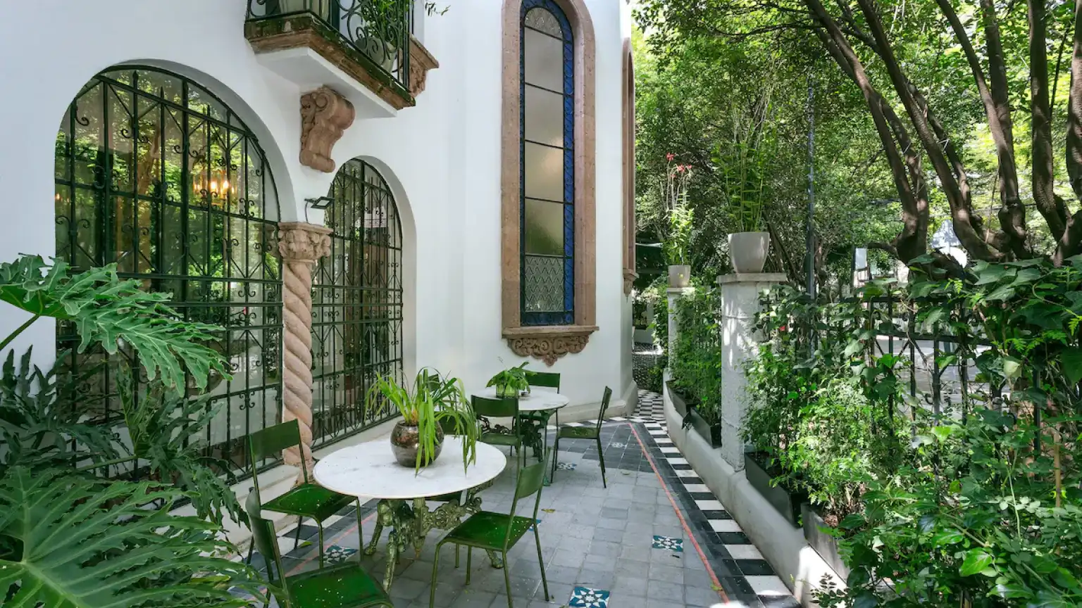 A side terrace beneath an arched stained-glass window and iron grilles, with green bistro tables on a chequered floor, among plants