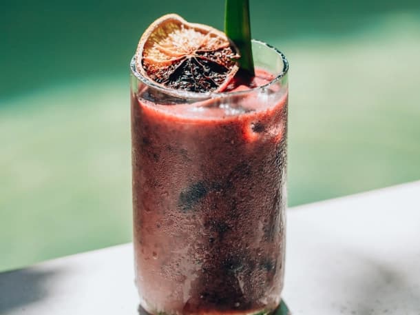 A red cocktail with a charred citrus wheel, beside the Orquidea pool