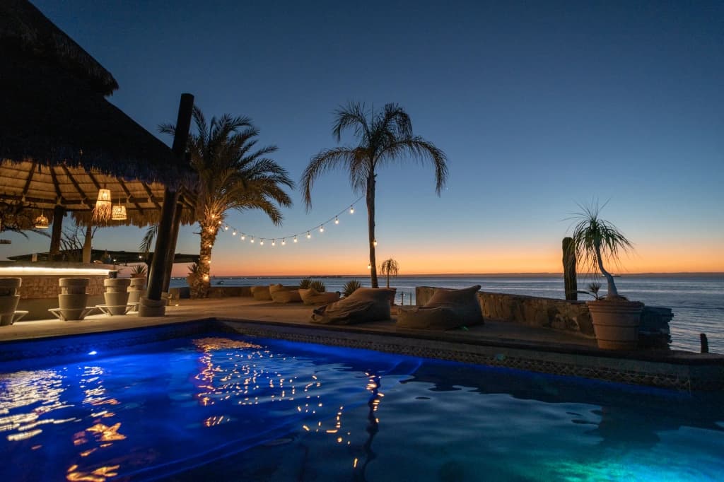 The pool at dusk, with string lights among the palm trees, the palapa bar lit up and the sea's horizon glowing orange