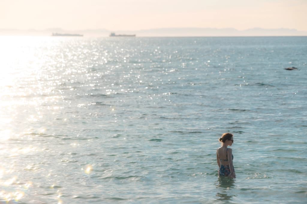 A guest standing in the calm sea, sunlight glinting on the water and the bay's mountains on the far shore