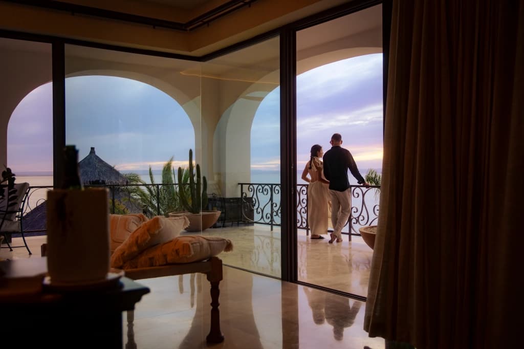 A couple on the balcony at sunset, looking out at the sea, seen through the arched glass doors from the room