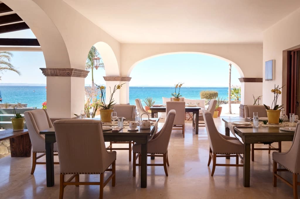 The dining terrace under the arcades, tables set for breakfast and the turquoise sea beyond, framed by the columns
