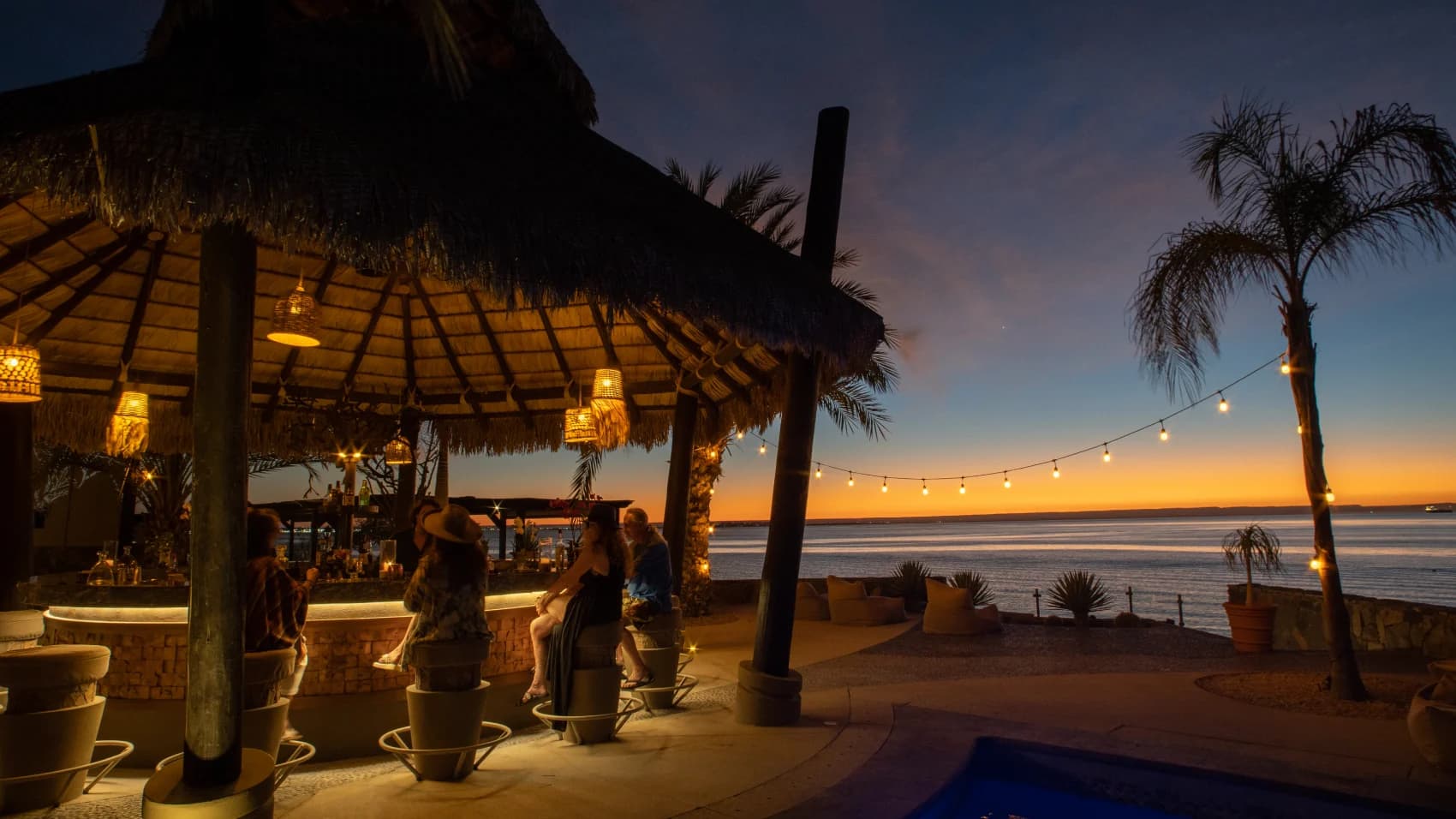 El Caimancito's palapa bar at dusk, guests at the stools and the sea glowing orange beyond