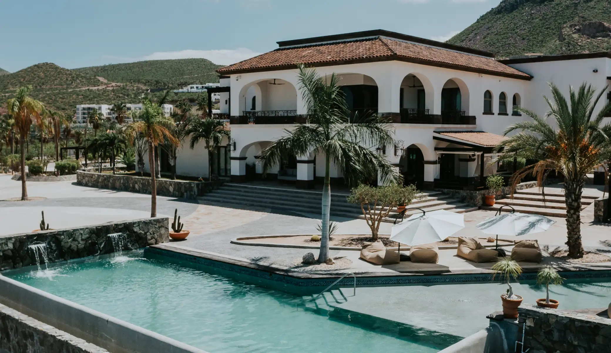 The two-storey house with its tiled roof and arcades, seen from the stone-edged pool with a small waterfall, among palm trees and the desert mountains beyond