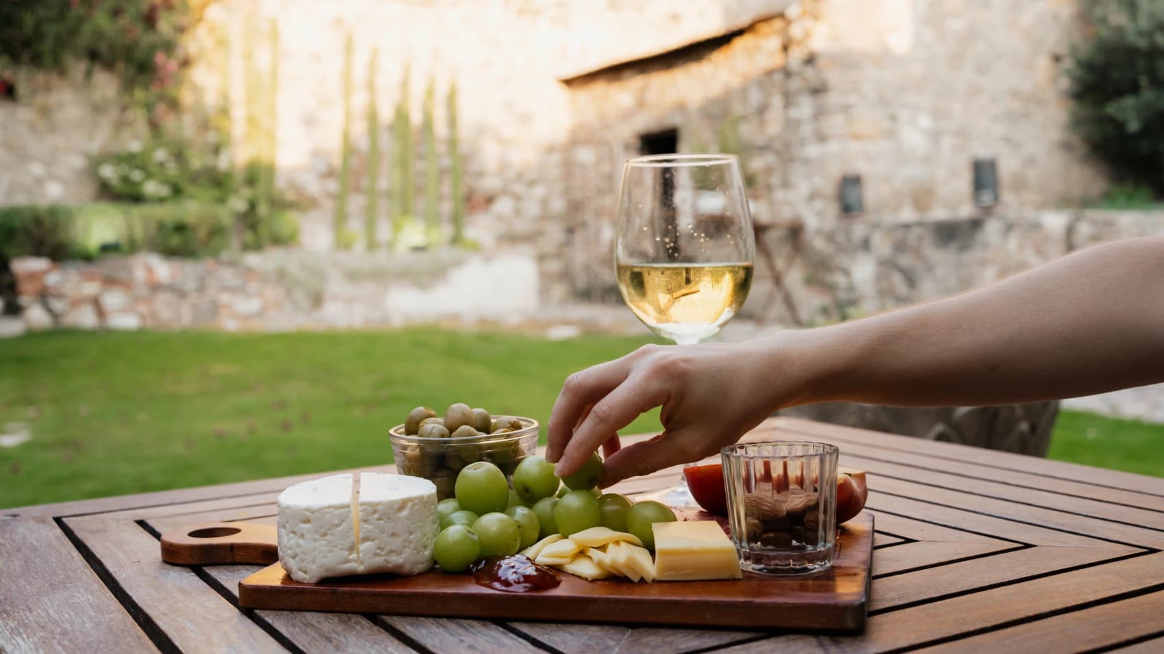 A board of cheese, grapes, olives and almonds on the garden table, a glass of white wine and the stone wall behind