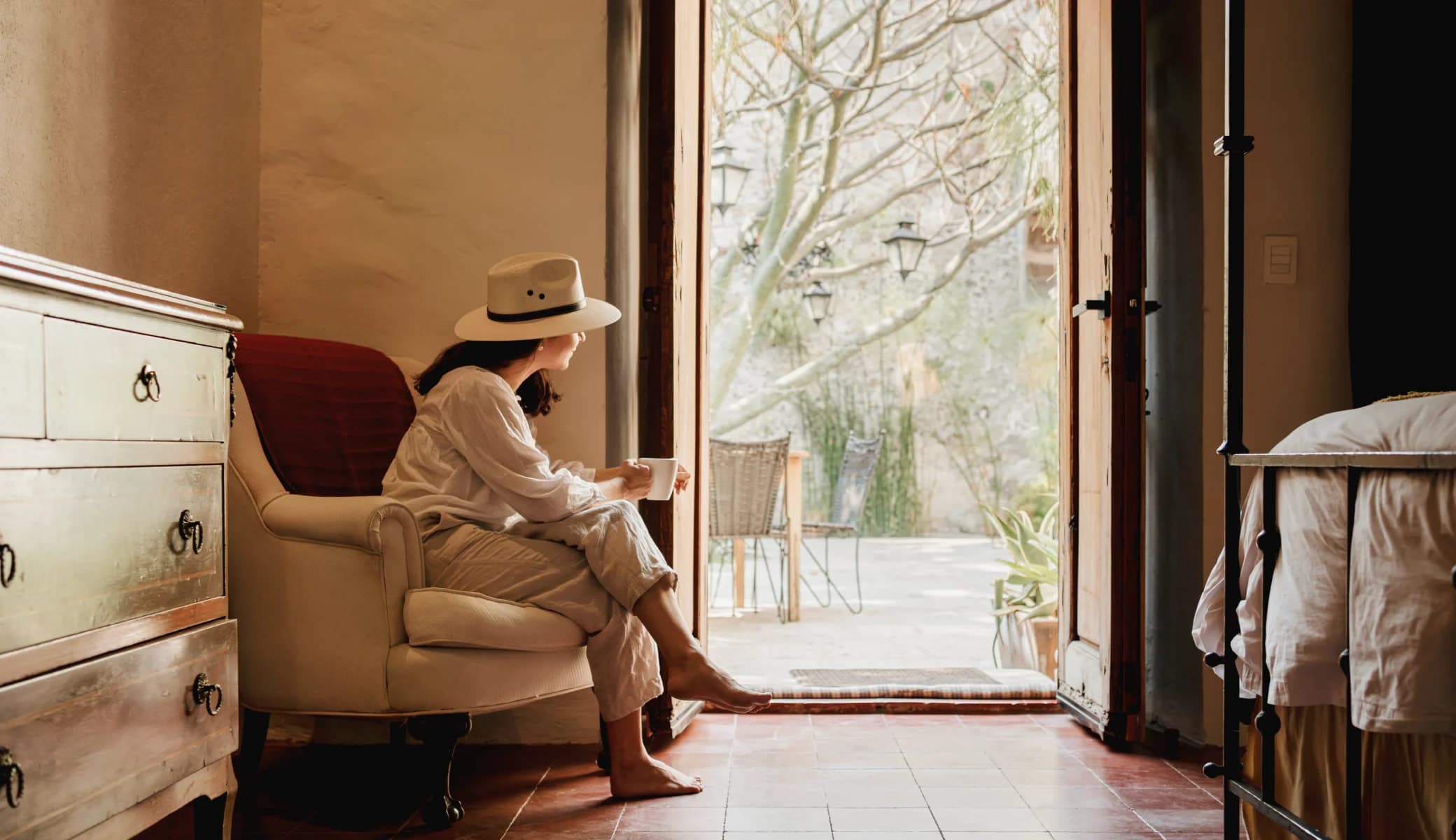 A guest in a straw hat has coffee seated in an armchair, looking out to the courtyard through the open wooden door