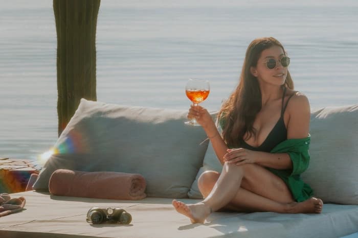 Someone in sunglasses on a daybed facing the sea, holding an aperitif, binoculars beside them