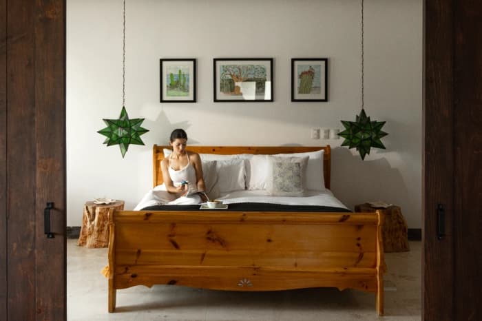 Una habitación con cama de madera tallada, lámparas verdes en forma de estrella y tres acuarelas de cactus sobre la cabecera; alguien desayuna sentado en la cama
