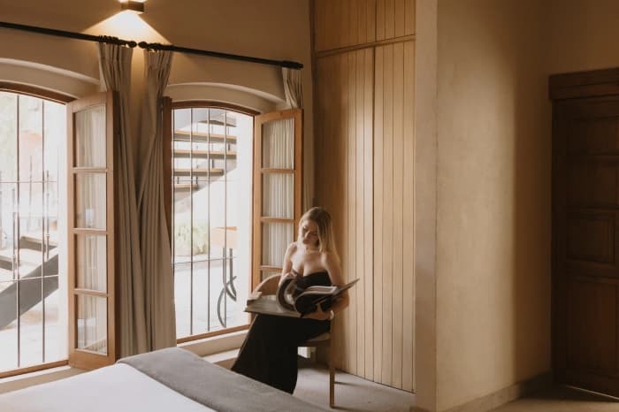 Someone reading a magazine by the room's arched windows, the linen curtains drawn back