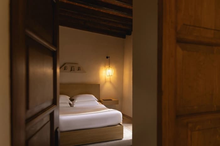 A room seen through the open wooden door: a bed with a pale wood headboard, an exposed beam and a lit wall lamp