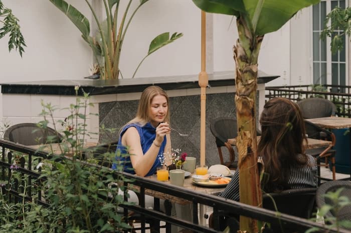 Two people at breakfast on the garden terrace, among banana plants and a palm, orange juice and flowers on the table