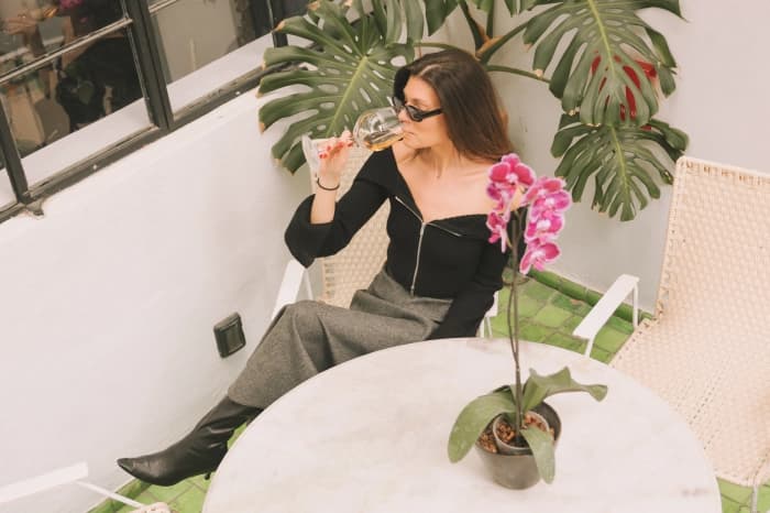 Alguien sentado en la terraza con una copa de vino blanco, junto a una mesa de mármol con una orquídea y una hoja de monstera detrás