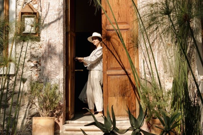 Alguien de lino claro y sombrero de ala en el umbral de la puerta de madera, entre papiros y agaves