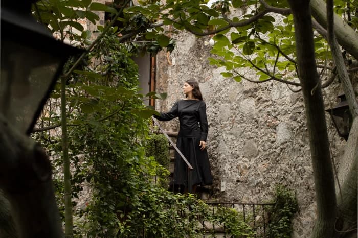 Alguien de vestido largo en una escalera de la casa, entre los muros de cantera y las ramas del patio