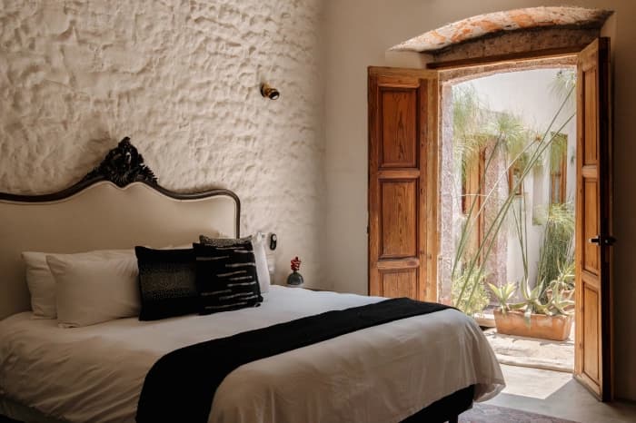 Una habitación de muros de piedra encalada, con cabecera tallada, cojines oscuros y la puerta de madera abierta al patio
