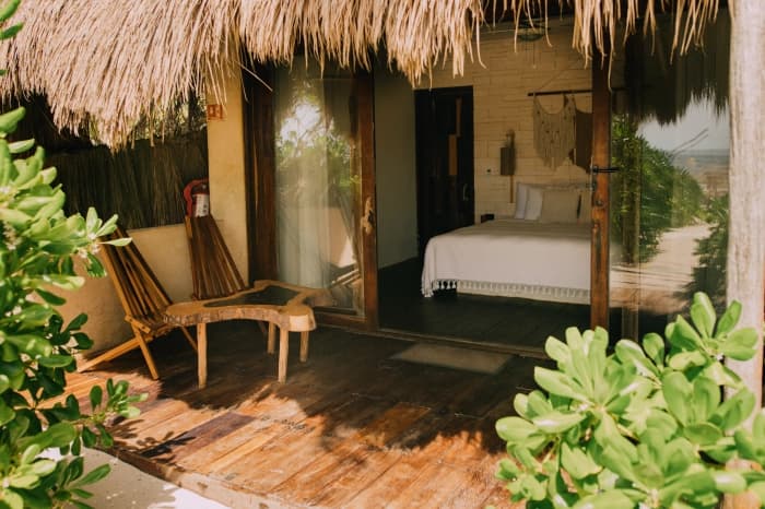 A room's wooden terrace under a palm roof, two lounge chairs and the bed beyond the glass doors