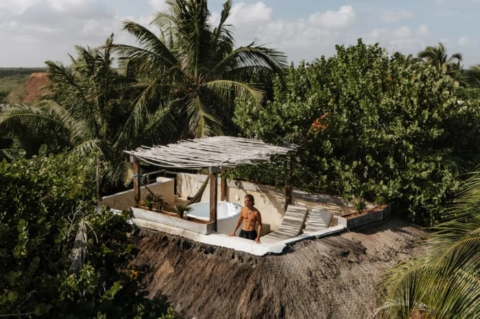 Aerial view of a clifftop terrace, with a tub and loungers under a timber pergola, ringed by palms