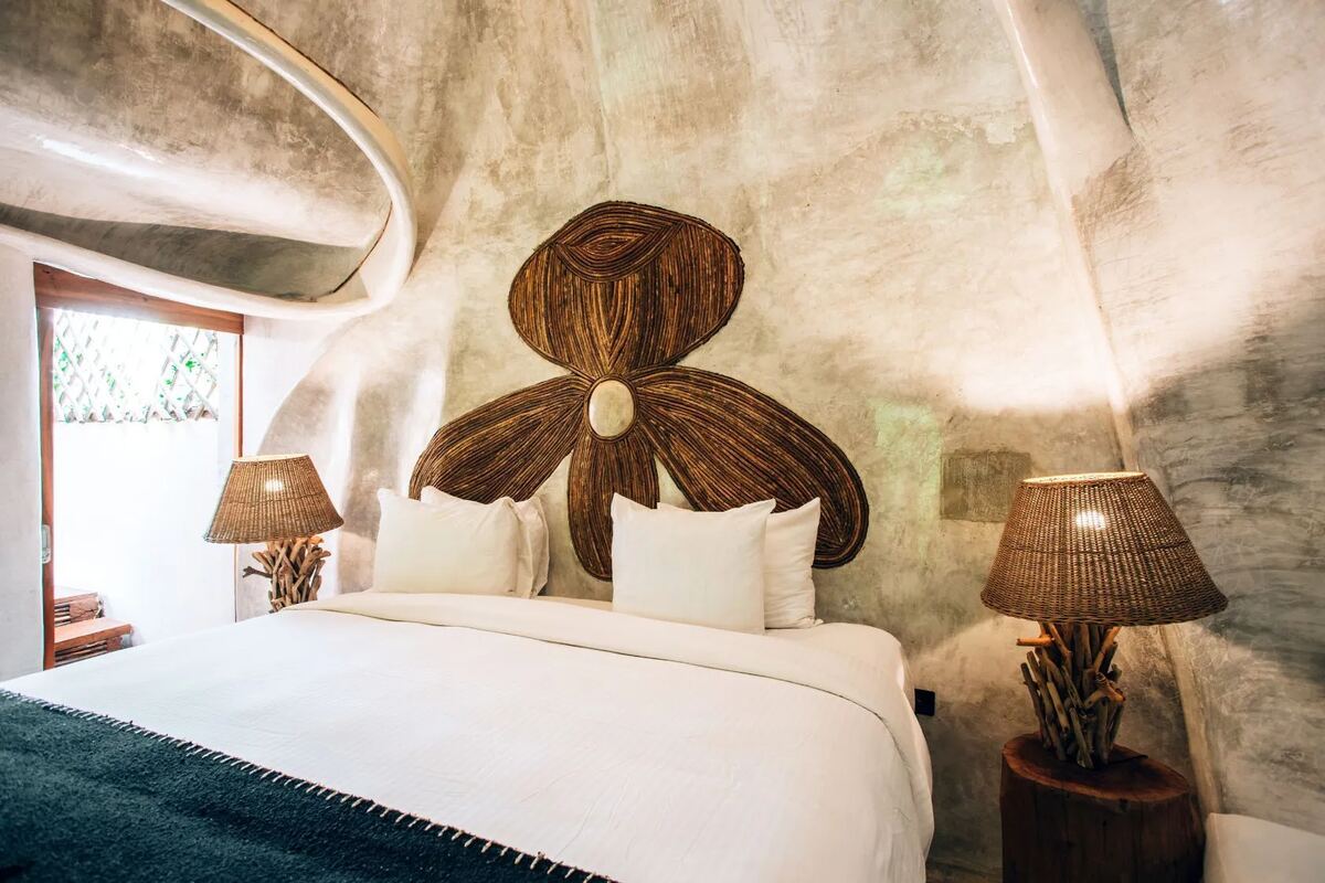 A studio with curved polished-concrete walls, a carved timber flower above the headboard and wicker lamps to either side