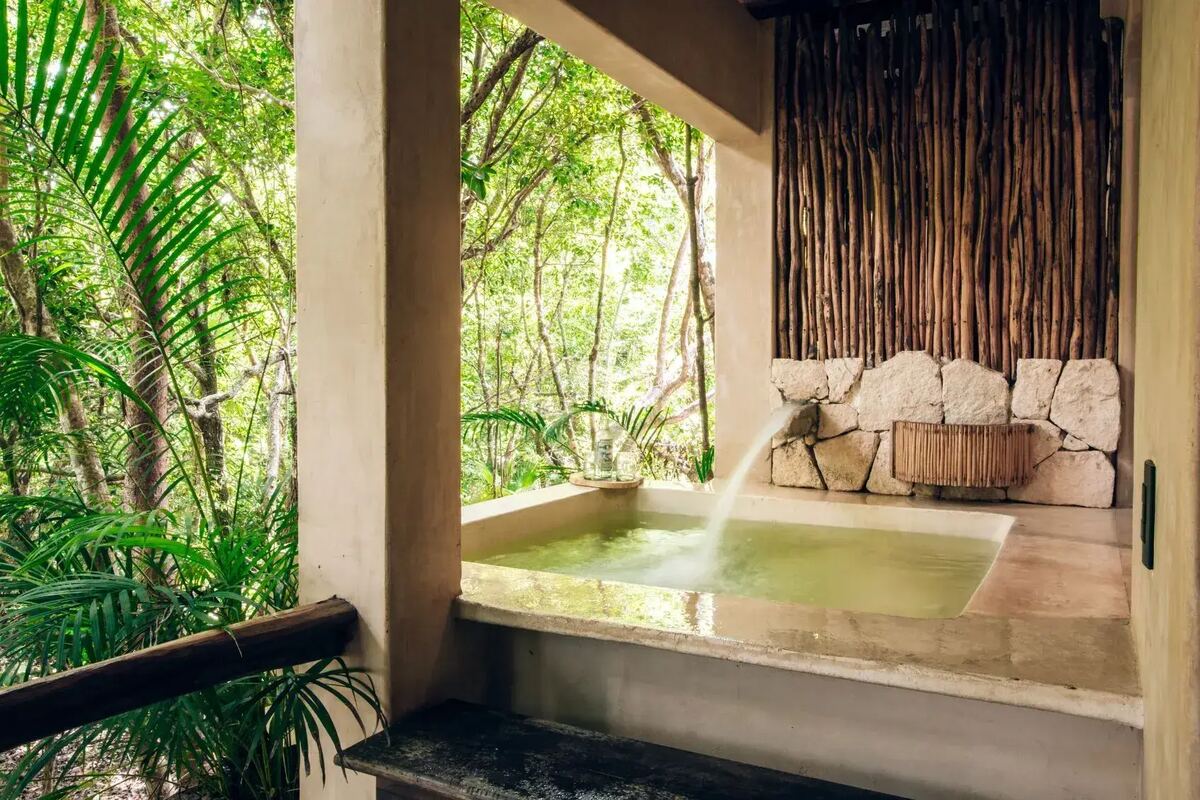 A stone bathtub filling in the open air, facing the jungle through a wall of timber posts