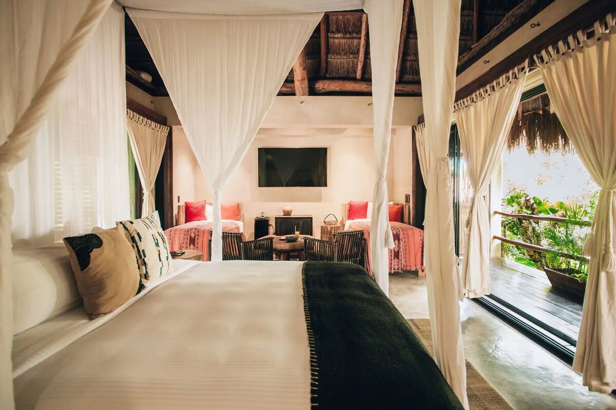 A master suite with a four-poster bed and white mosquito netting under the palm roof, and a sitting area beyond