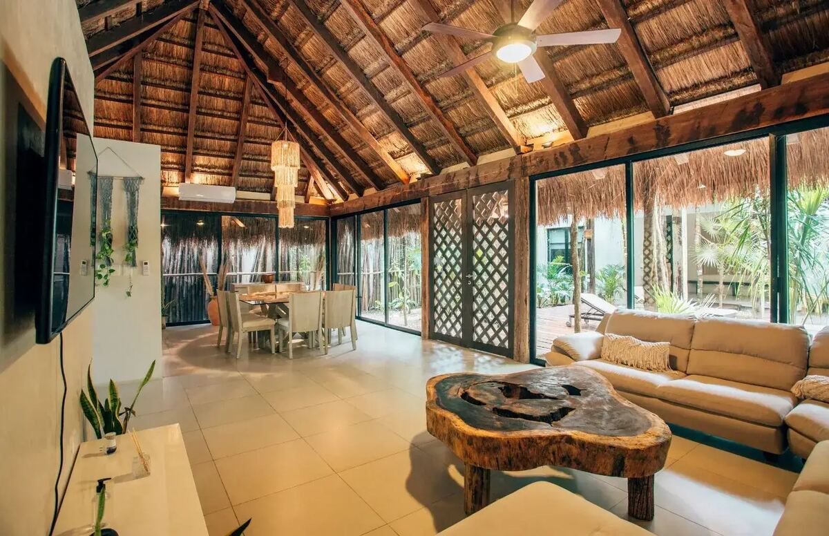The villa's living room under its pitched palm roof, with its own dining table and glass onto the jungle on both sides