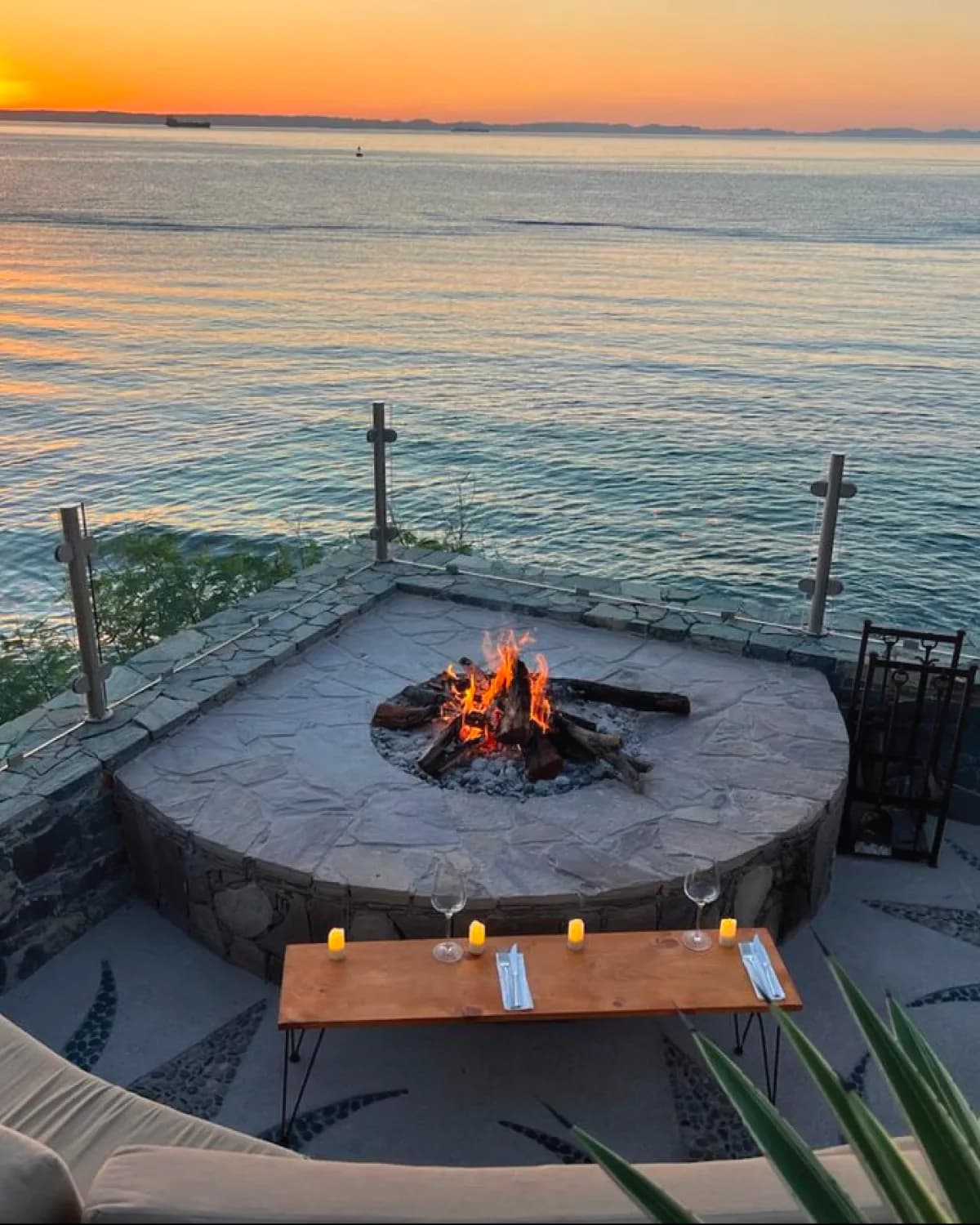 Stone terrace above the water with a lit fire pit, a low table with glasses and candles, and the sun setting over the bay