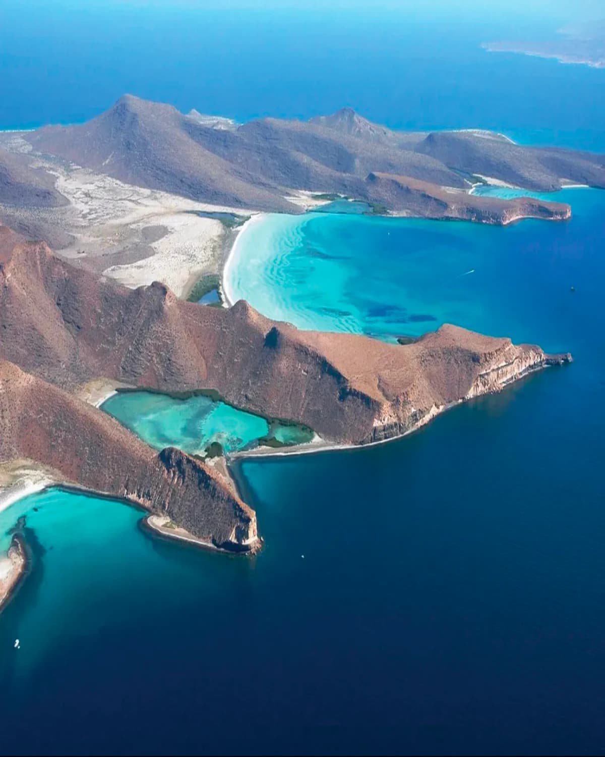Aerial view of the island: brown rocky ridges dropping to the sea, with turquoise coves and deep blue water around them