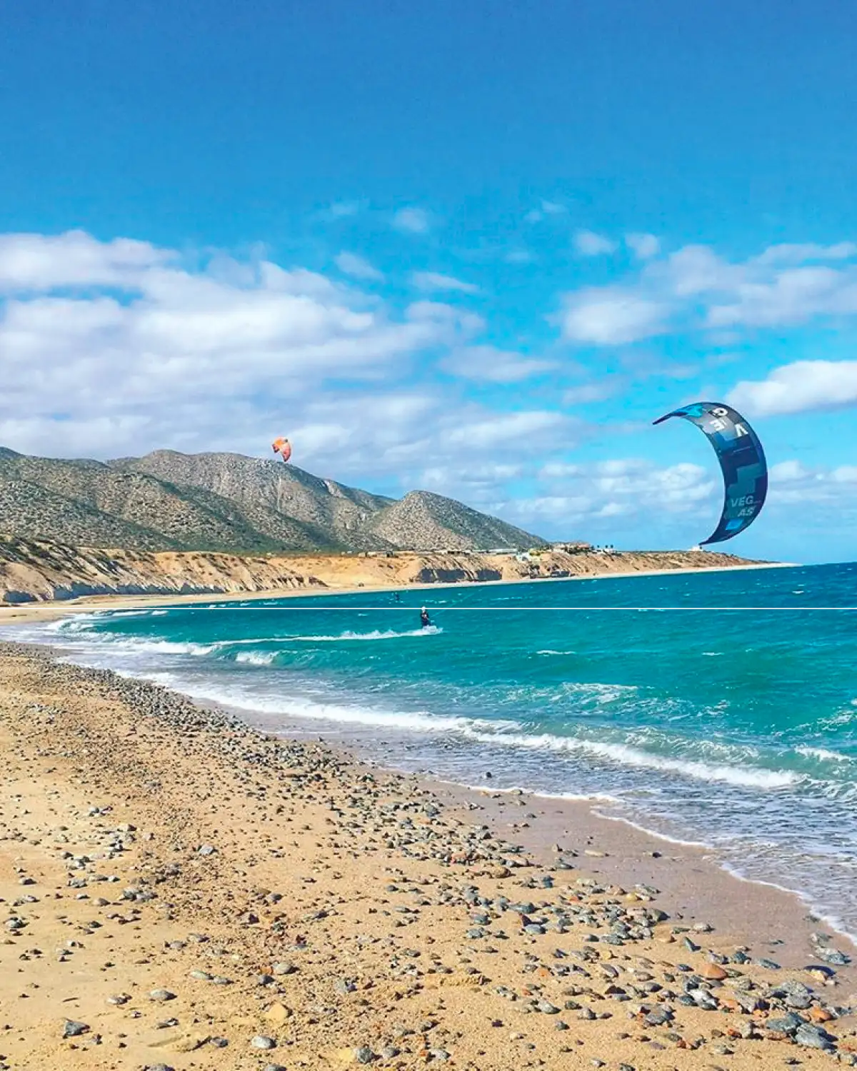 Sand-and-pebble beach with turquoise water, a kitesurfer riding and kites in the air, against the mountain range