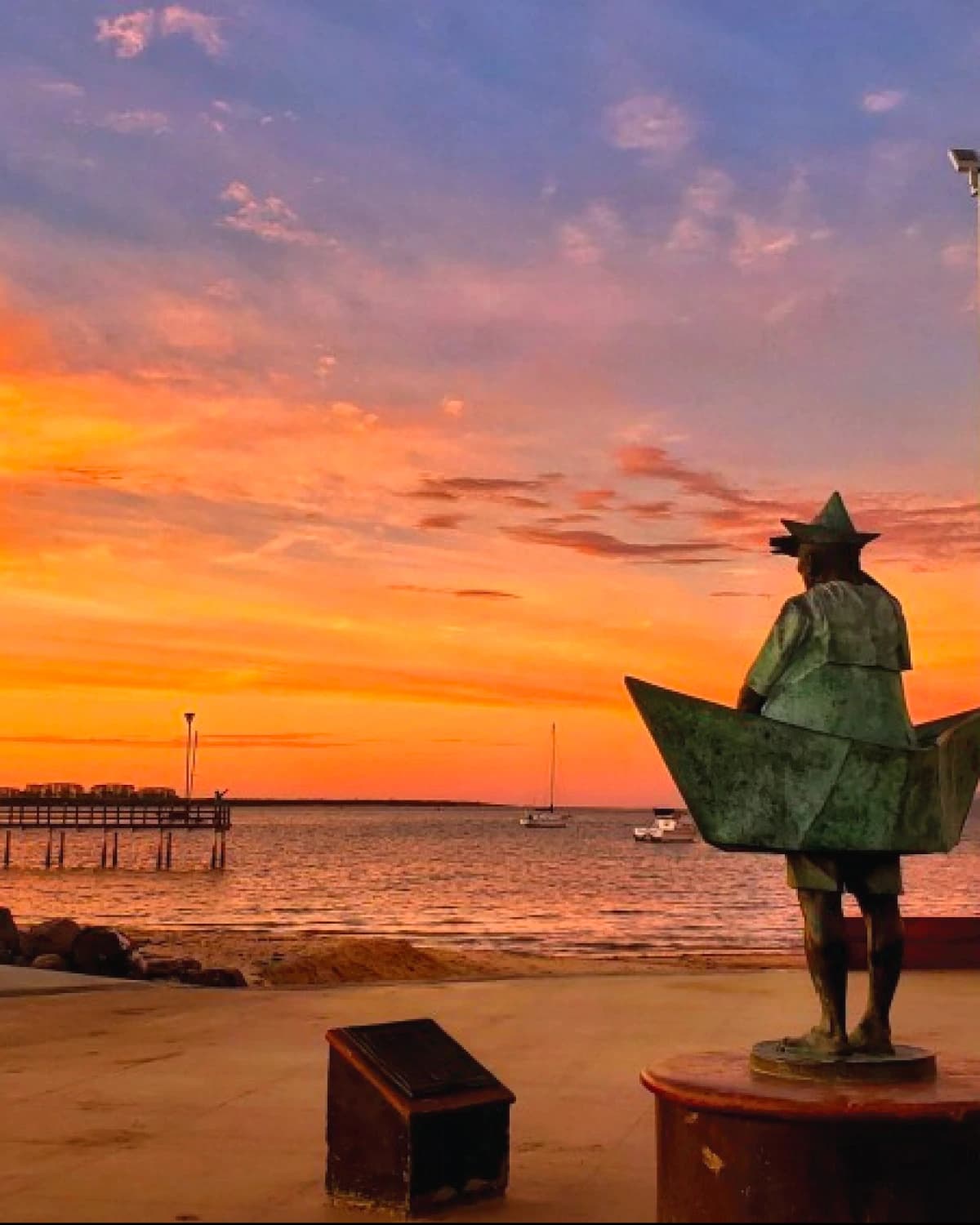 The La Paz malecón at sunset: the bronze sculpture of a figure inside a paper boat, with the pier and a few sailboats on the orange water
