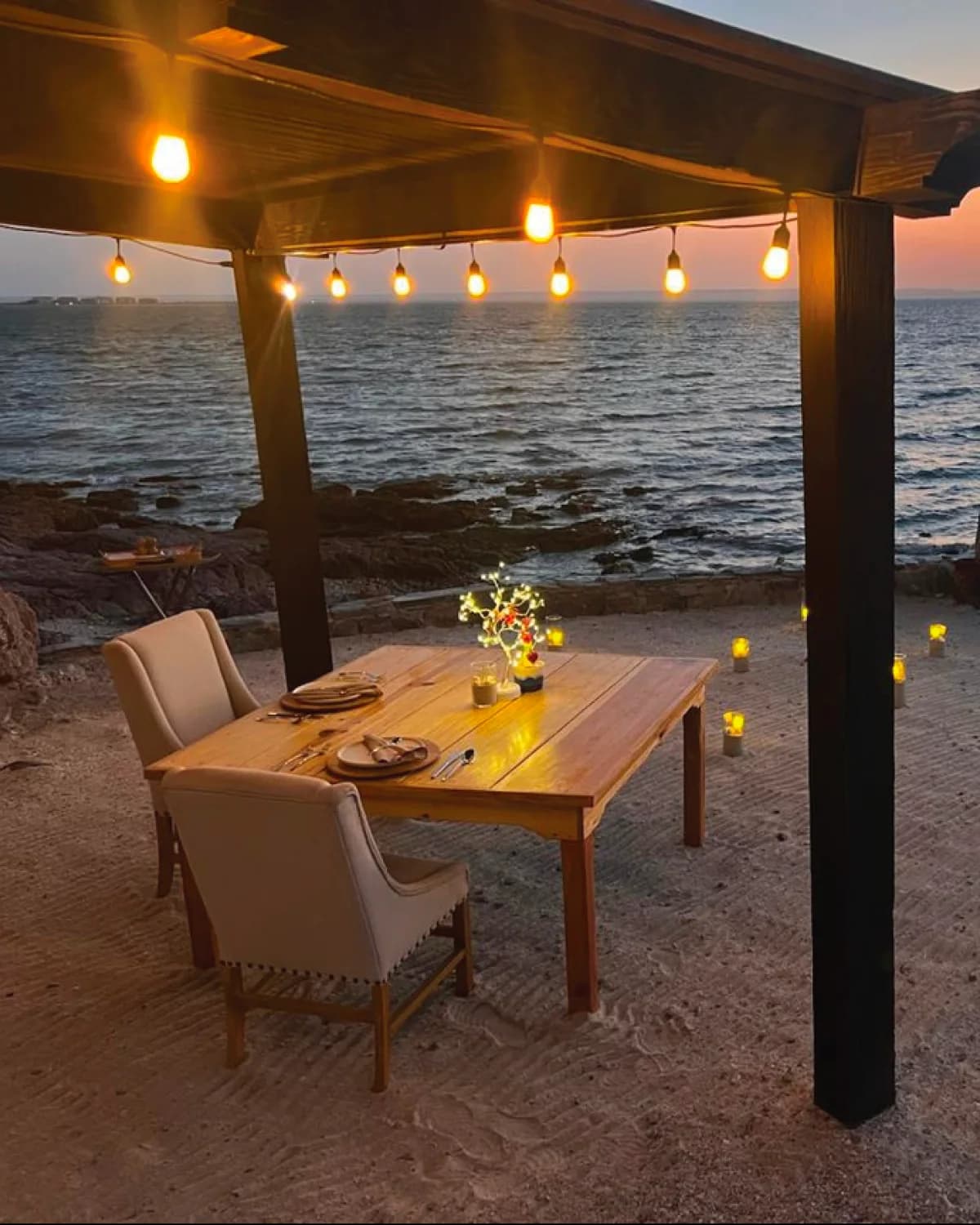 Wooden table set for two under a pergola strung with bulbs, on the sand facing the sea at dusk, with lit candle lanterns around it