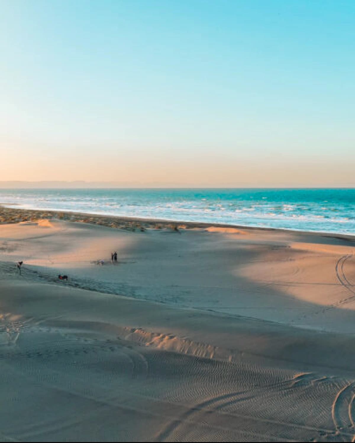 Sand dunes at sunset, wind ripples across the surface, the sea beyond and a few small figures in the distance
