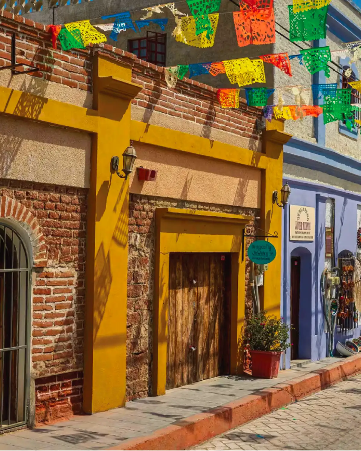 Street with colorful papel picado strung overhead, a yellow facade with a wooden door and a purple house with a jewelry shop next door