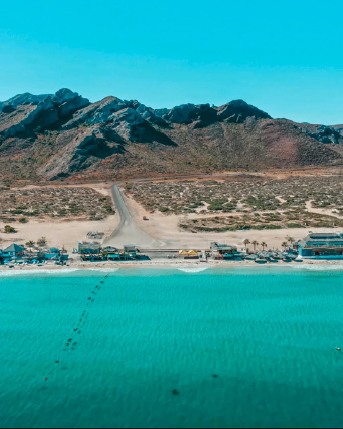 Aerial view of a turquoise-water beach with palapas and beach clubs along the shore, and desert hills behind