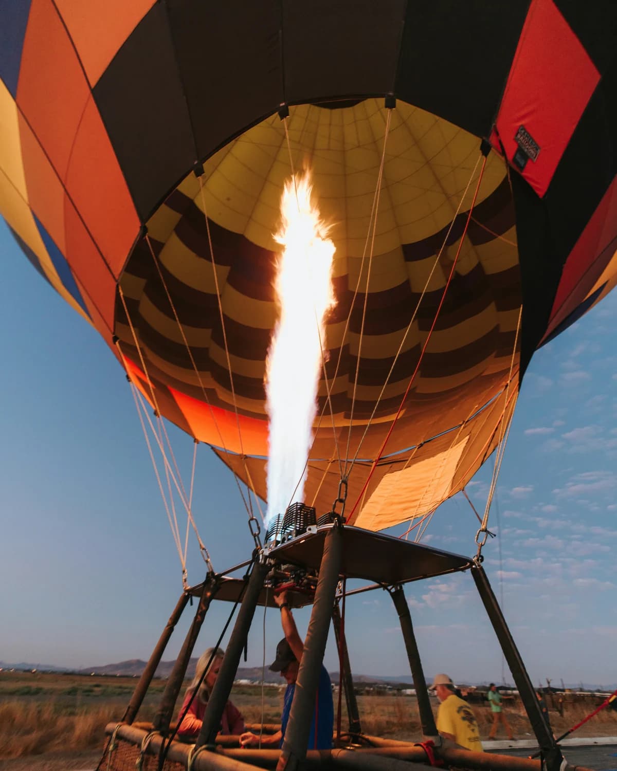 An orange, black and red hot air balloon inflating with the burner's flame lit, guides preparing the basket at dawn