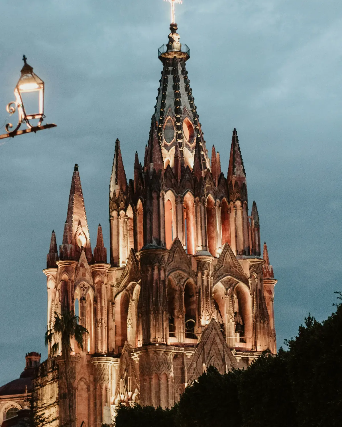 The spires of the Parroquia de San Miguel Arcángel at dusk, lit, with a street lantern in the foreground