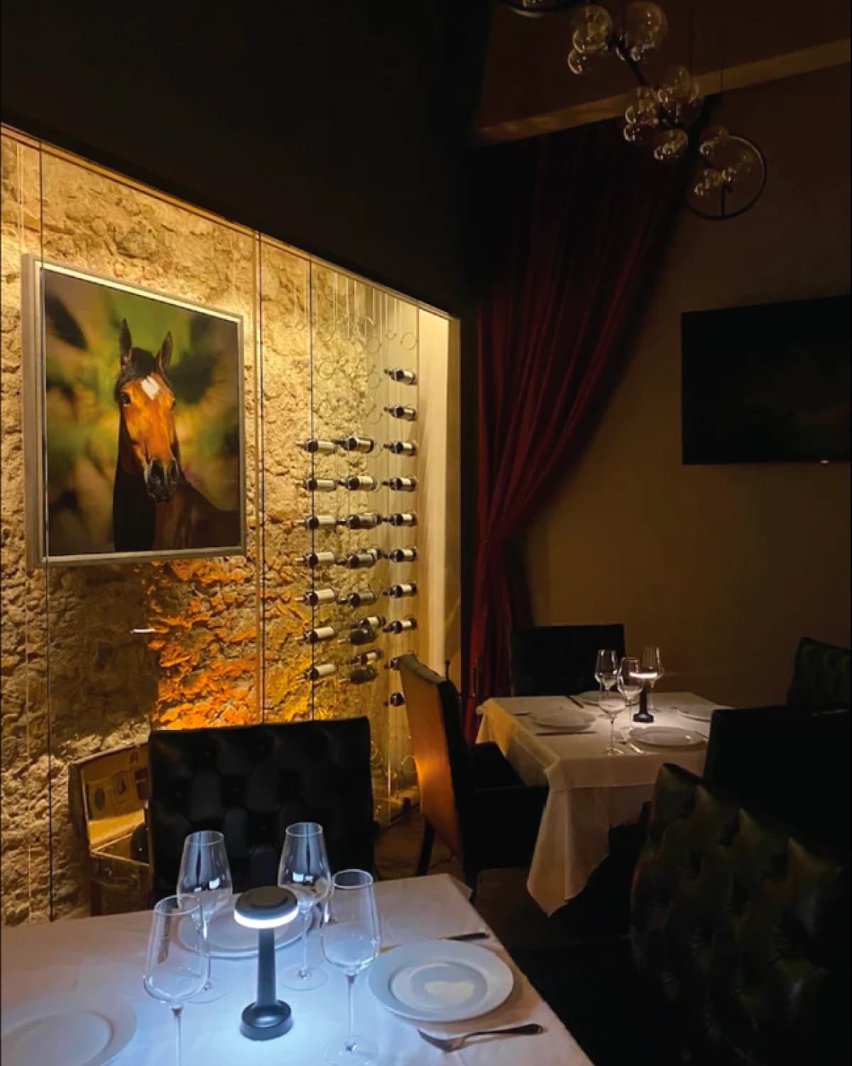 Dimly lit dining room: white-clothed tables set with glasses, tufted chairs, a stone wall with a horse portrait and a vertical wine rack