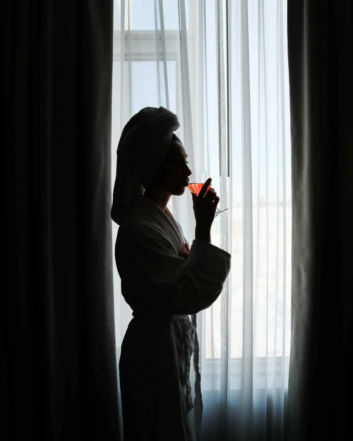 Silhouette of a guest in a robe and towel turban, sipping a drink by a window with sheer curtains