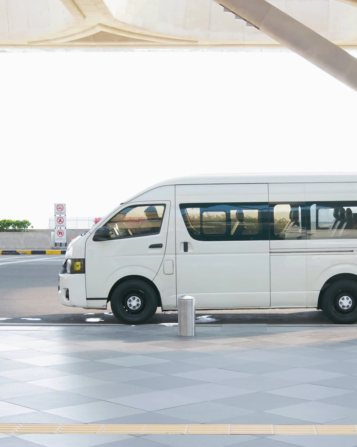 A white transfer van, seen from the side, parked by the curb under the terminal canopy