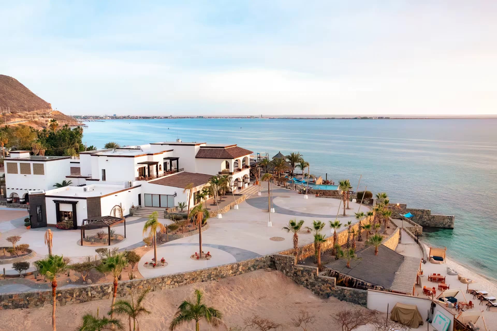 The house seen from above at sunset: tiled roofs and white arcades, palm trees and the pool on the point, with the bay of La Paz and the city beyond