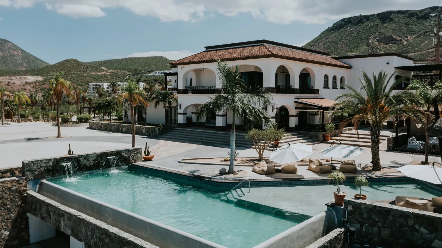 An elevated view of the facade and the infinity pool with its waterfall edge, among palm trees and loungers under white umbrellas