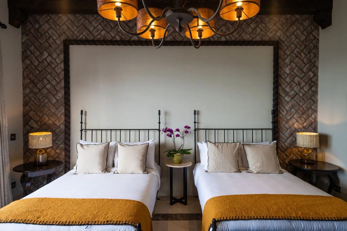 Room with two wrought-iron beds, a herringbone brick wall, a shaded chandelier and an orchid