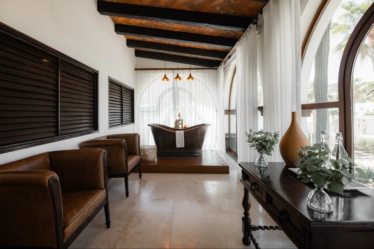 Suite with a freestanding copper tub on a timber deck, facing an arched window with white curtains