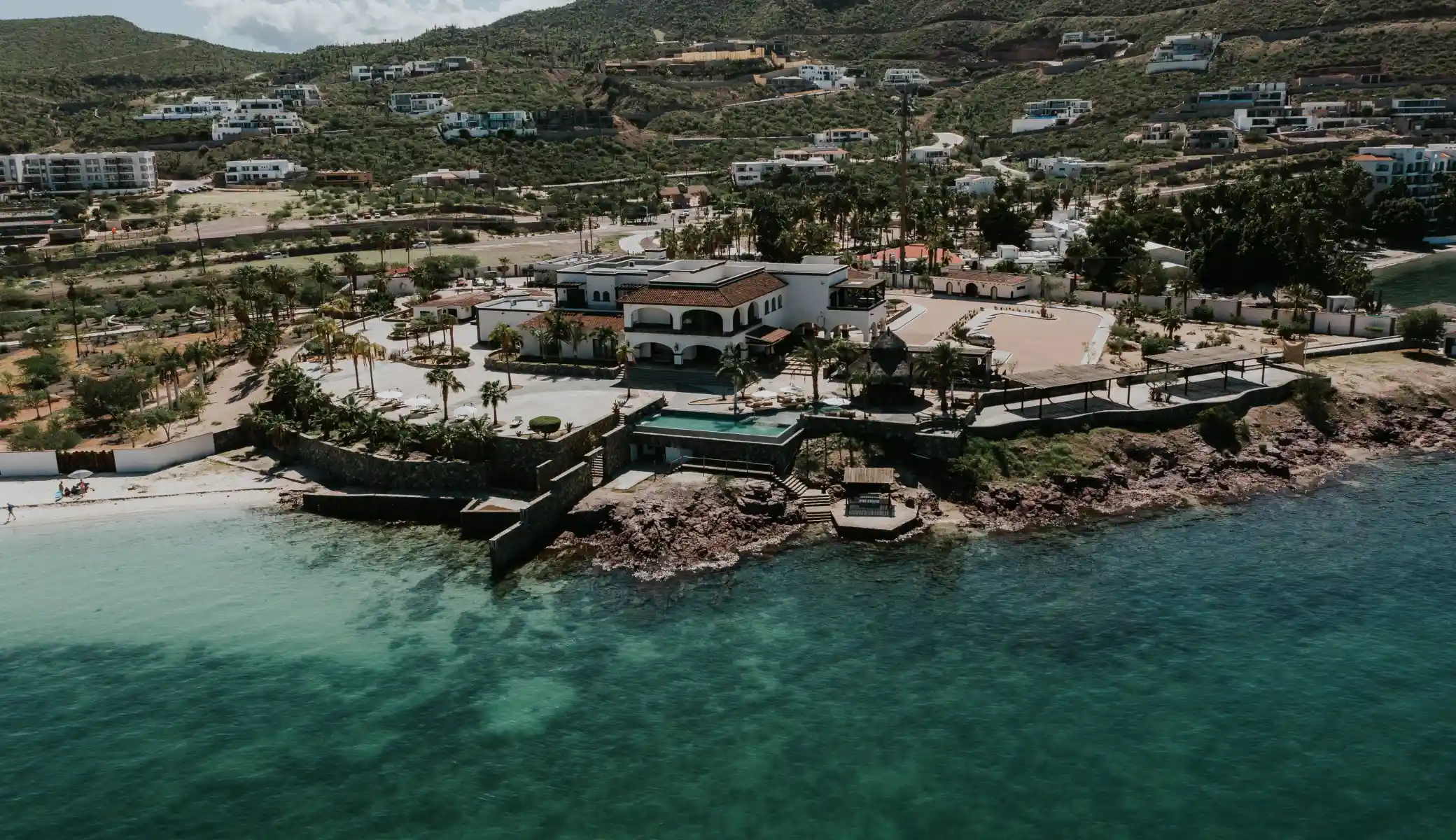 Aerial view of Orchid House Baja on the rocky point: the house, the pool and the beach club's palapa, between the turquoise sea and the hills of La Paz