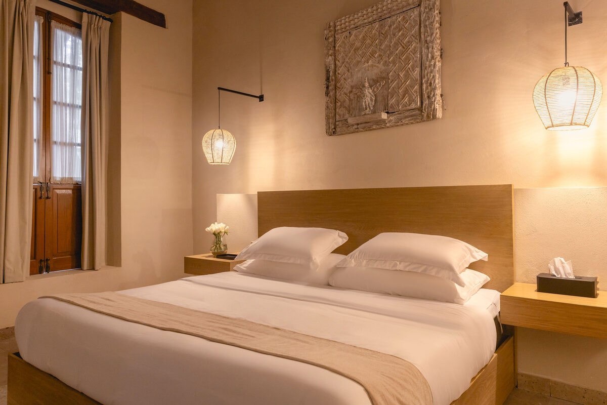 Recámara con cama king bajo un relieve tallado enmarcado, con viga de madera a la vista y puertas de madera al costado