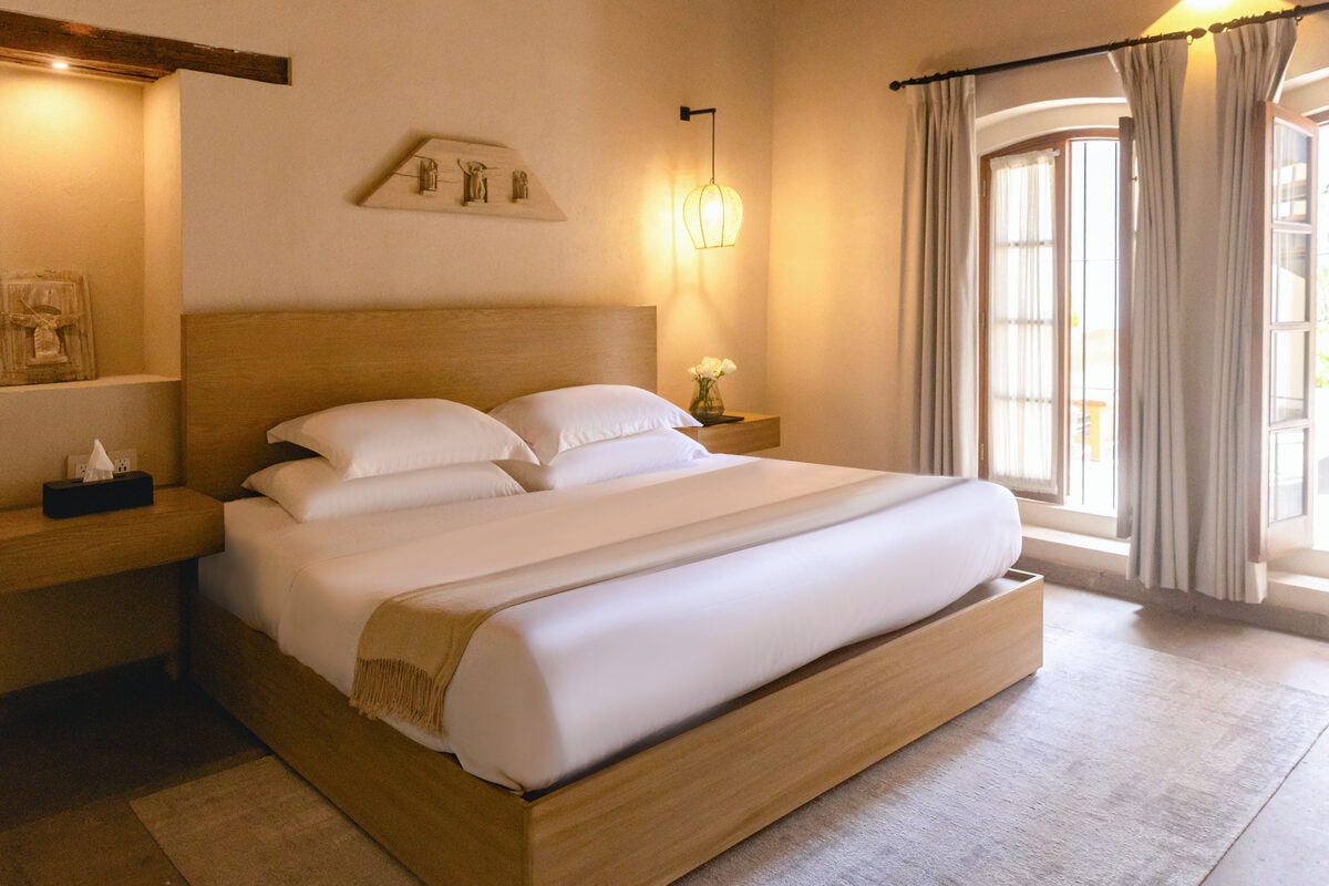 Recámara con cama king junto a un ventanal con cortinas, bajo una viga de madera