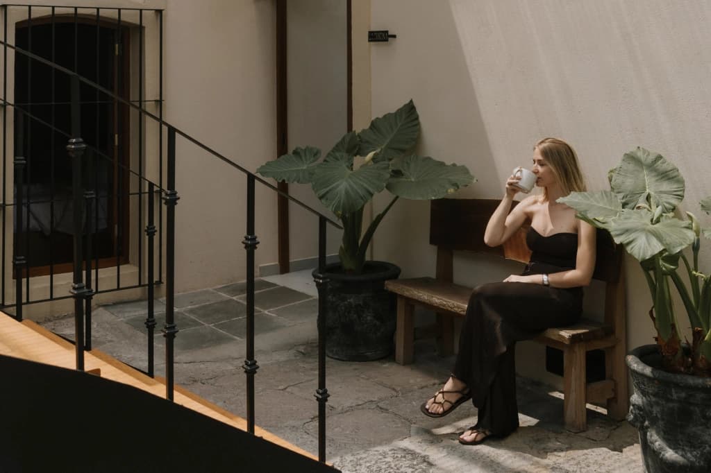 Una huésped toma café sentada en una banca de madera, junto a plantas de hoja grande y el barandal de herrería de la escalera