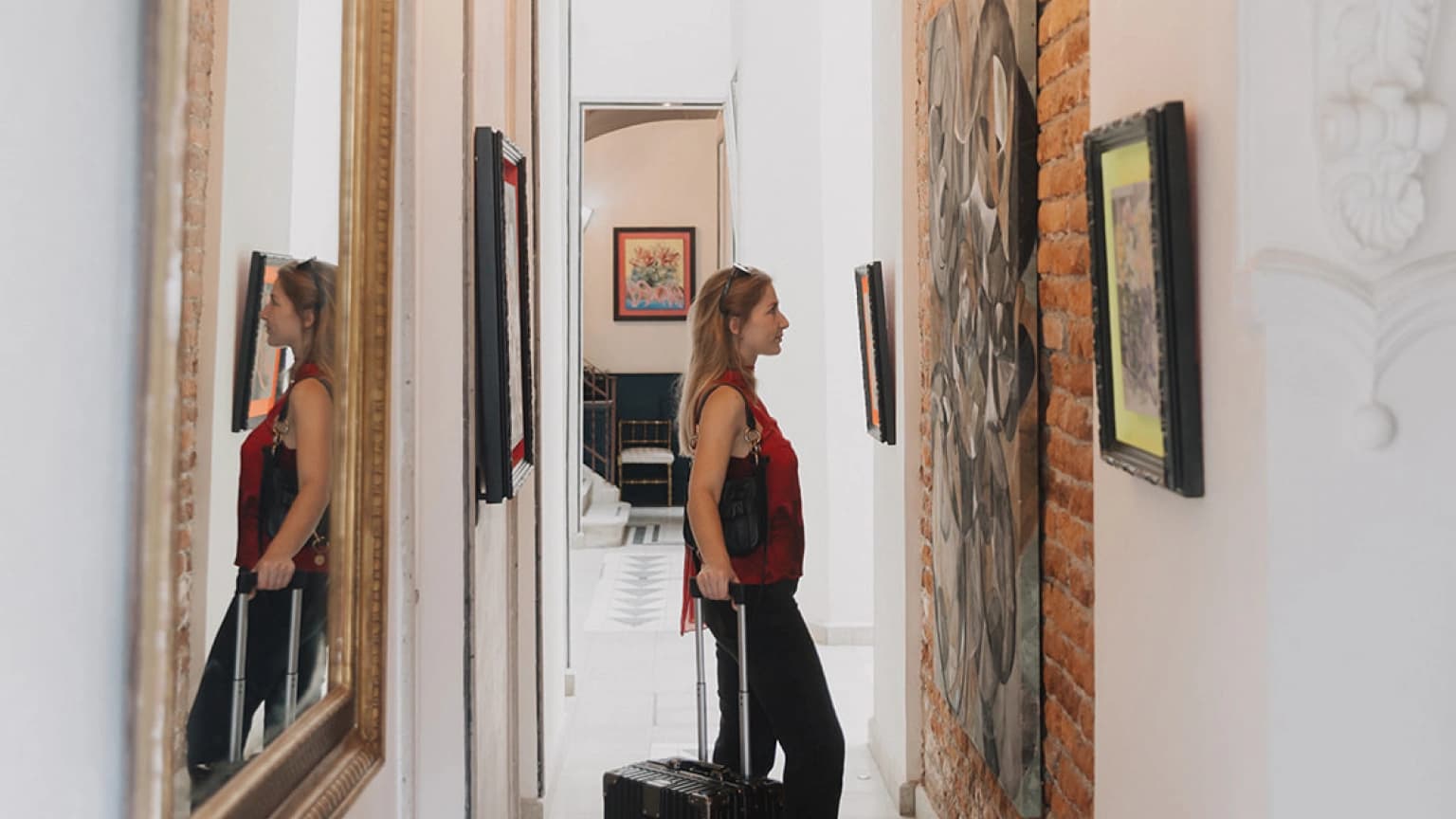 A guest with a suitcase in a brick corridor with framed art and a gilded mirror